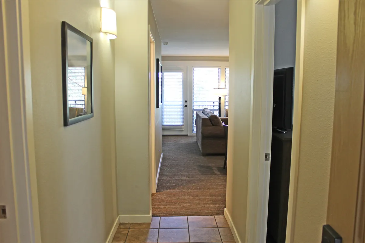 A view down a hallway into a sunlit living room with access to the balcony through glass doors. The image captures the transition from indoor comfort to the promise of fresh mountain air and scenic views.