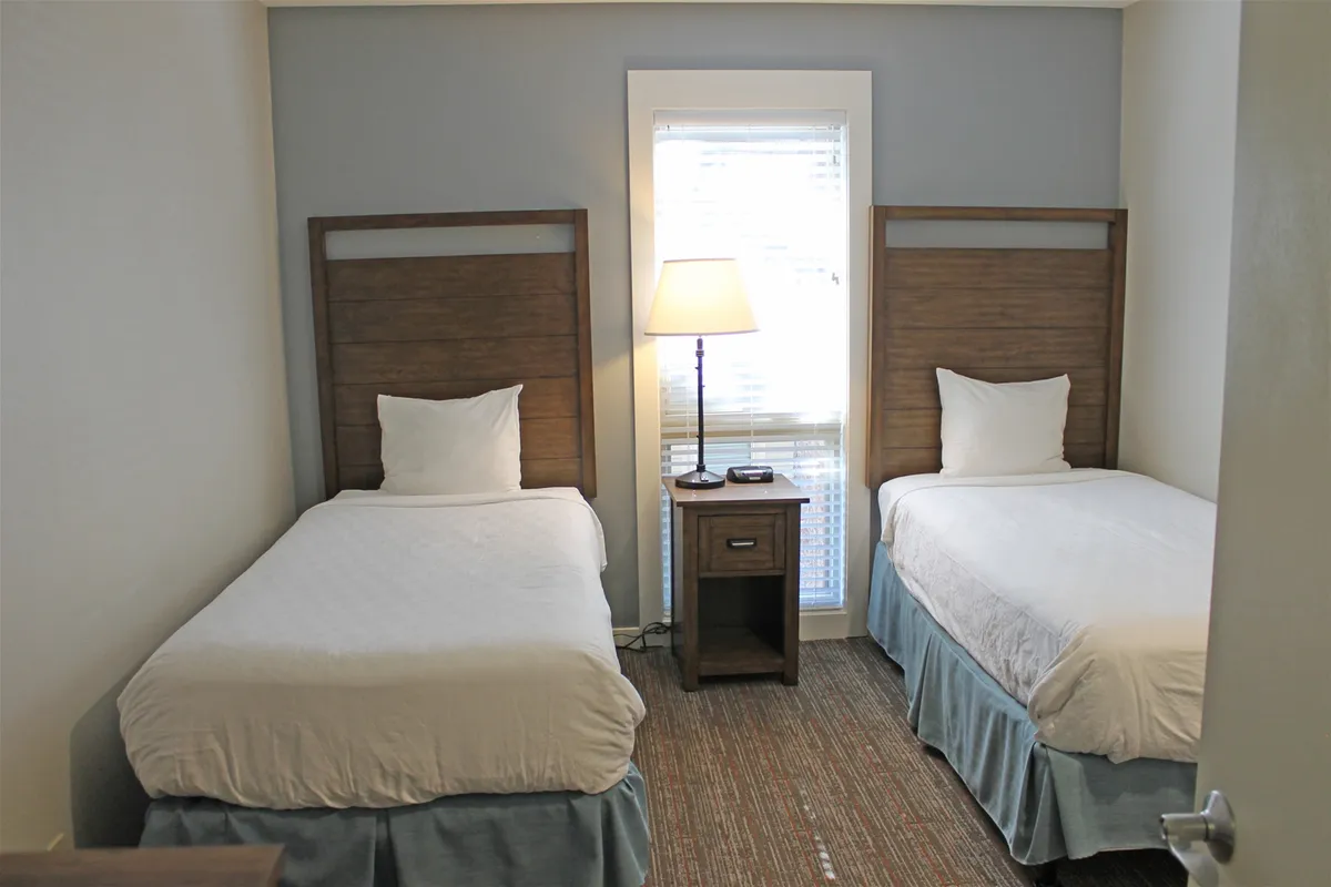 A bright, cozy room featuring twin beds separated by a nightstand with a lamp, a large window letting in natural light, and welcoming touches that suggest flexibility and comfort for children, friends, or couples.