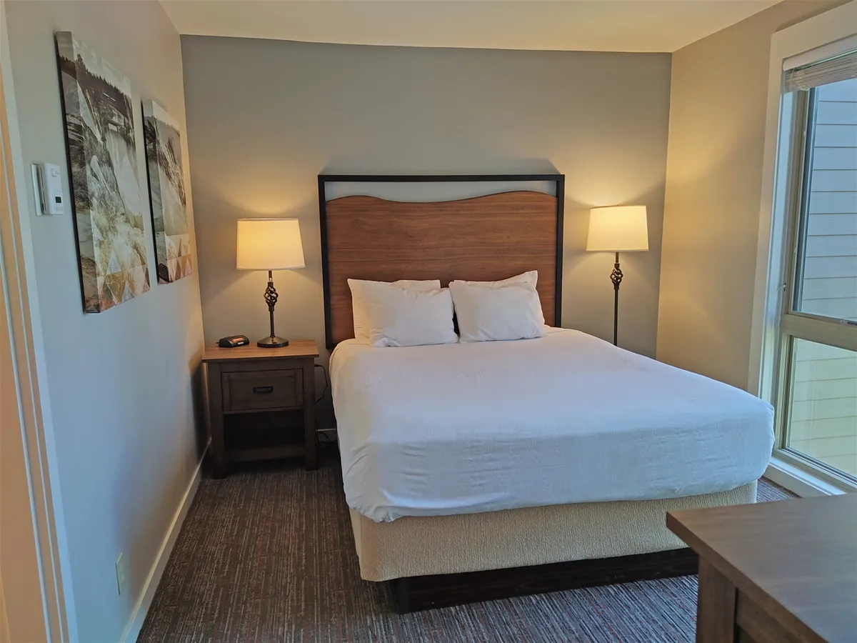 A quiet bedroom with a queen bed adorned in soft linens, flanked by nightstands and lamps, and complemented by tasteful artwork. The space feels inviting, cozy, and private, suggesting a restful escape.