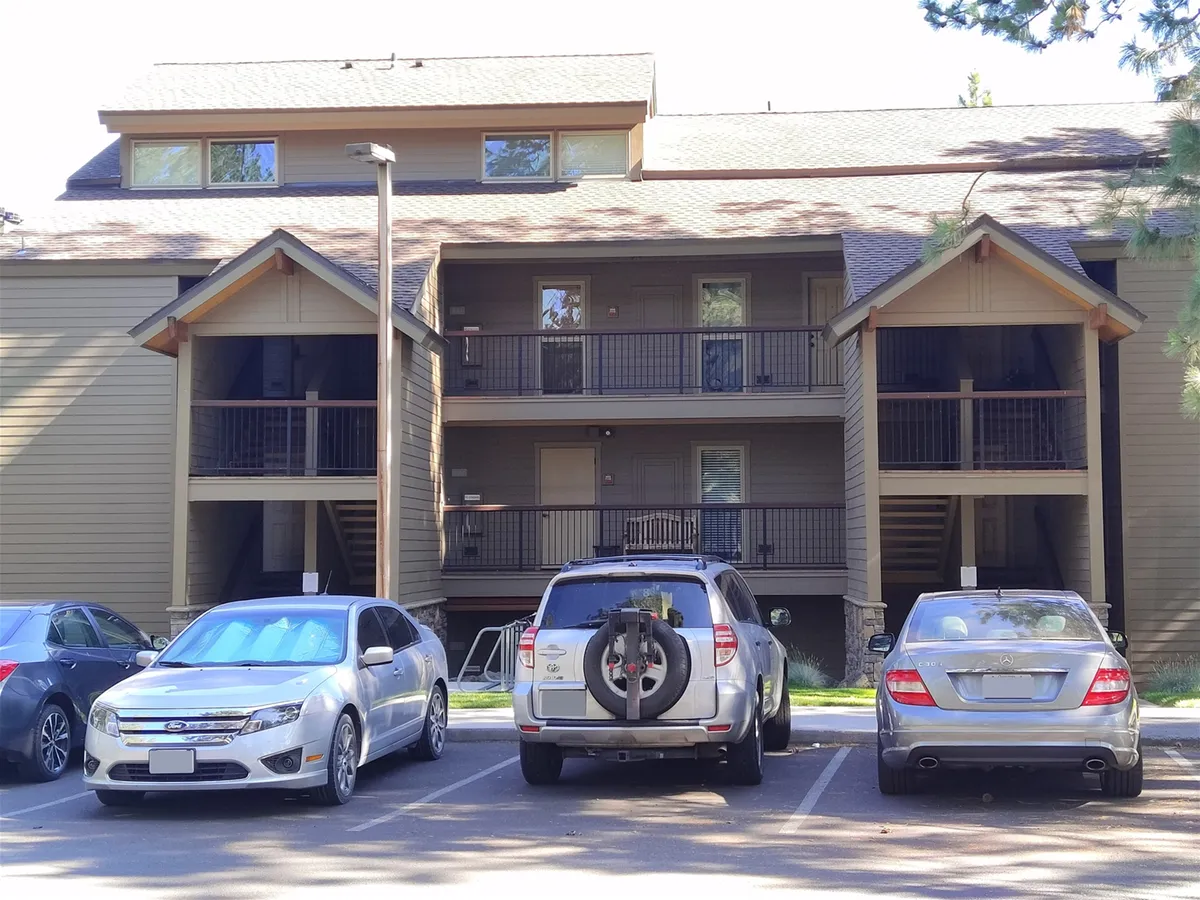 A two-story apartment building is framed by balconies and parked cars, with a friendly, open atmosphere. The communal outdoor space invites families and groups to gather before heading out to Mt. Bachelor or after a day of winter fun.
