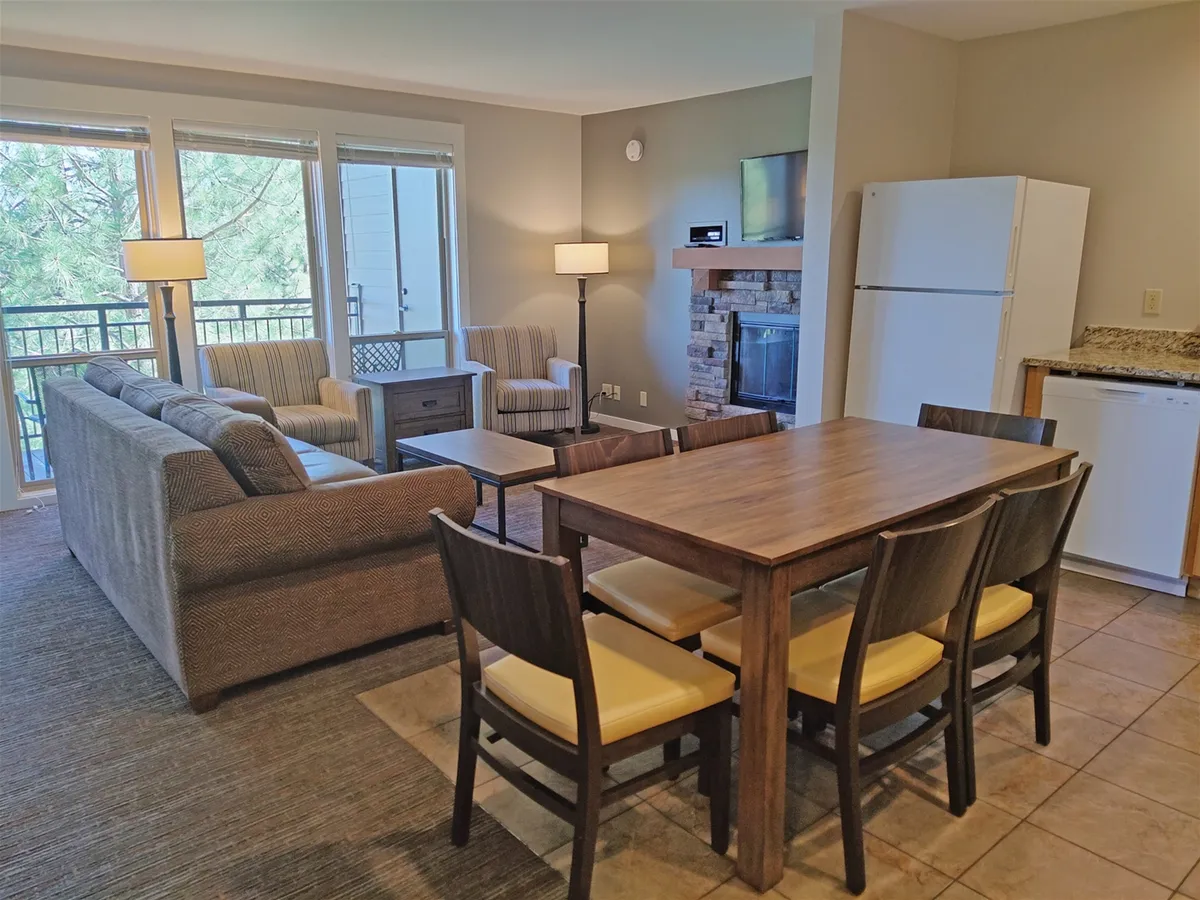 The image showcases a welcoming living and dining area with a plush sofa facing a crackling fireplace, a spacious dining table perfect for gathering, and oversized windows flooding the space with natural light and offering scenic views. The inviting atmosphere suggests cozy evenings, shared meals, and opportunities to take in the mountain landscape from the comfort of the condo.