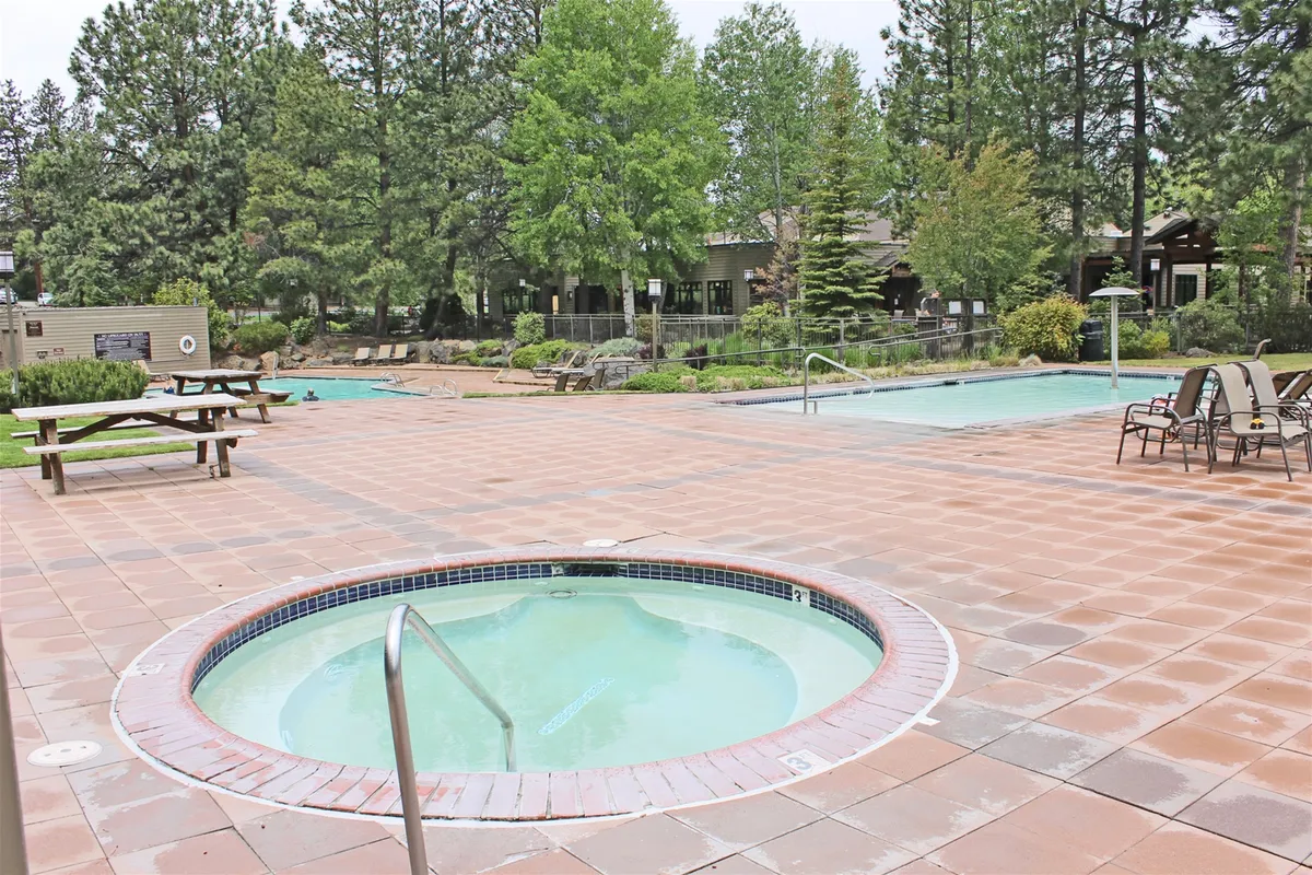 A vibrant outdoor scene featuring a hot tub and swimming pool surrounded by lush greenery and patio seating. The space is designed for relaxation and group gatherings, with ample room for socializing, lounging, and enjoying the fresh air.