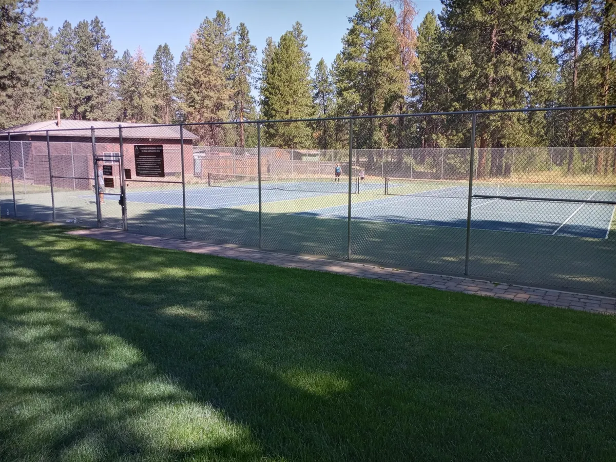 An exterior shot featuring well-maintained tennis courts surrounded by lush green grass and a fence, with a building visible in the background. The scene conveys the active and communal spirit of the resort's outdoor amenities.