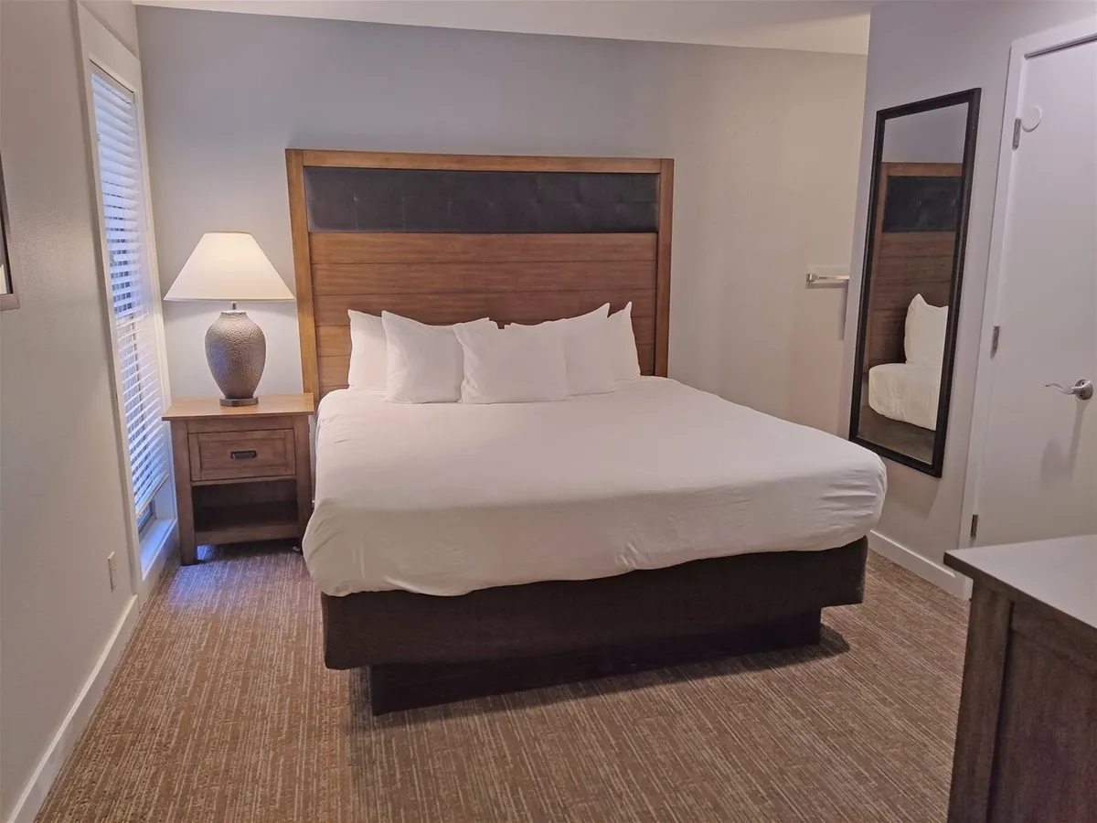 A spacious king-sized bed sits against a serene backdrop, accompanied by a nightstand with a lamp and a full-length mirror. The room exudes comfort and tranquility, bathed in soft, natural light, suggesting a restful night's sleep and peaceful mornings.